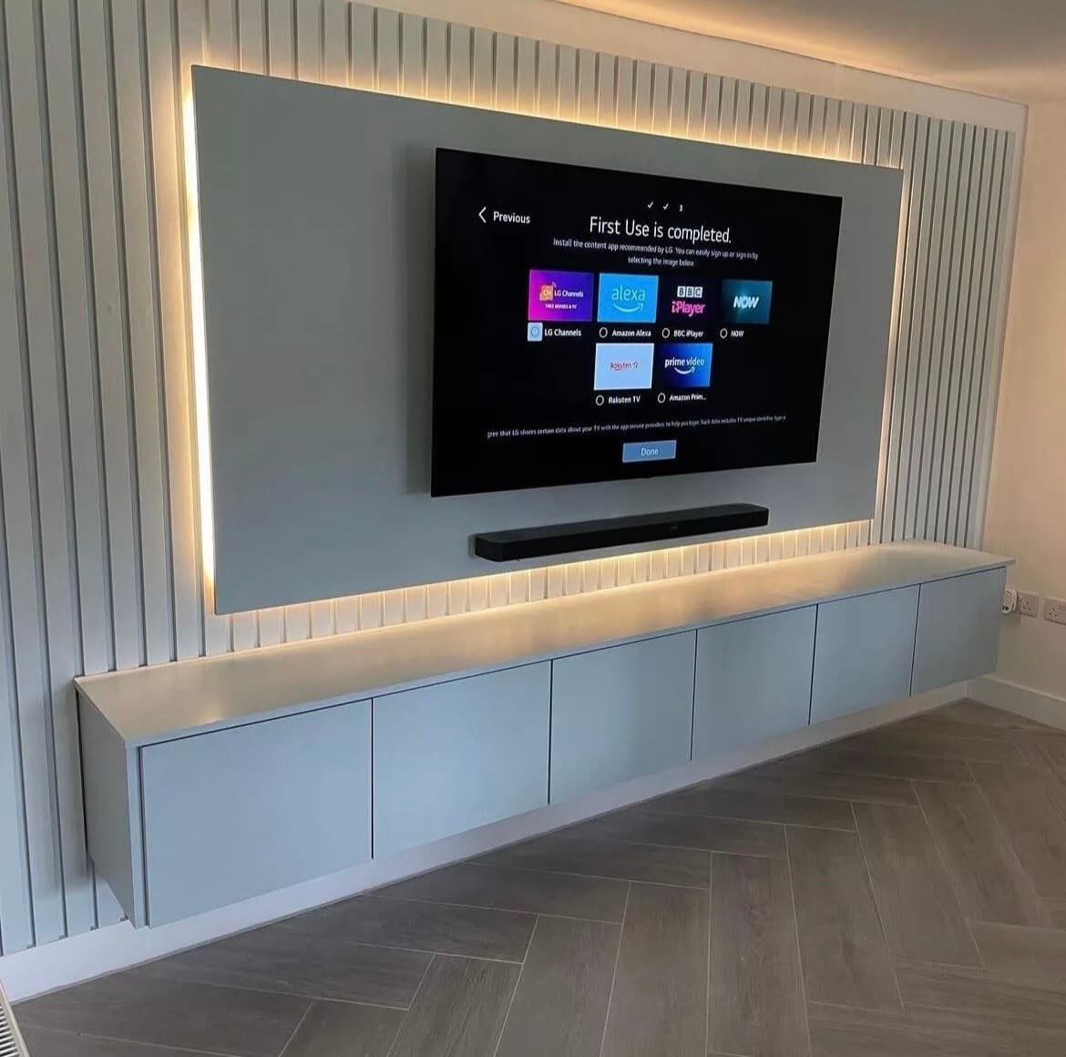 Modern wall-mounted TV with LED backlighting above a floating white cabinet on a slatted wall.